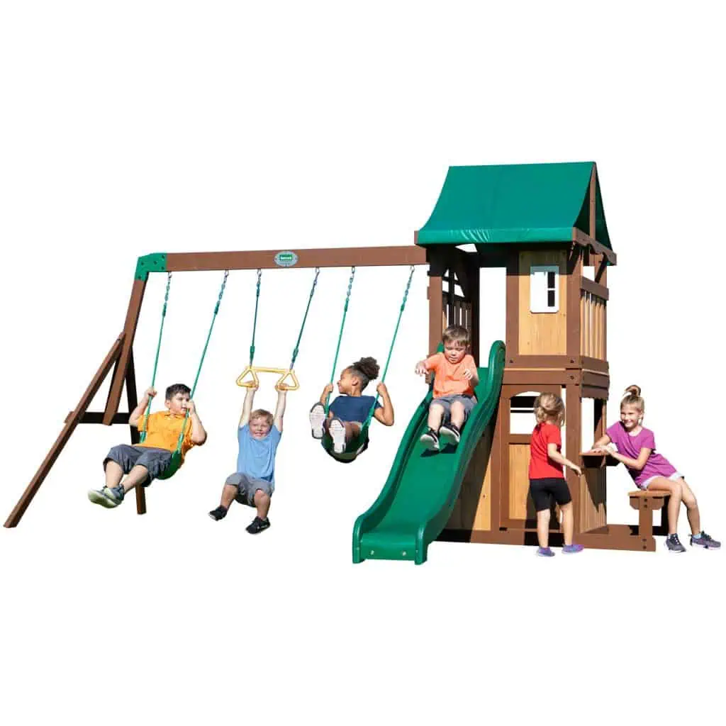 A large group of children are playing on a multi-featured wooden playset with forest green accents, isolated against a solid white background. On the left, three children are using the swing beam: two children are on green belt swings while a boy in a blue shirt hangs from a yellow trapeze bar in the center. To the right, a boy in an orange shirt slides down a wavy green plastic slide. The main two-story tower features a green fabric gabled roof, tan siding, and a white window on the upper level. On the far right, two girls are interacting at a built-in wooden picnic table attached to the side of the tower. The structure is built with dark brown wood framing and green metal supports.