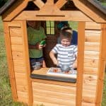 A close-up view through the large side opening of a wooden playhouse shows two children playing inside. In the foreground, a young boy in a striped t-shirt is "cooking" with a toy pot on a white play stovetop that features orange burner details. Behind him, a young girl in a yellow shirt watches while holding a small green and yellow toy. The playhouse is made of light-colored wood panels, and the scene is brightly lit by natural sunlight, with a grassy field visible in the background.