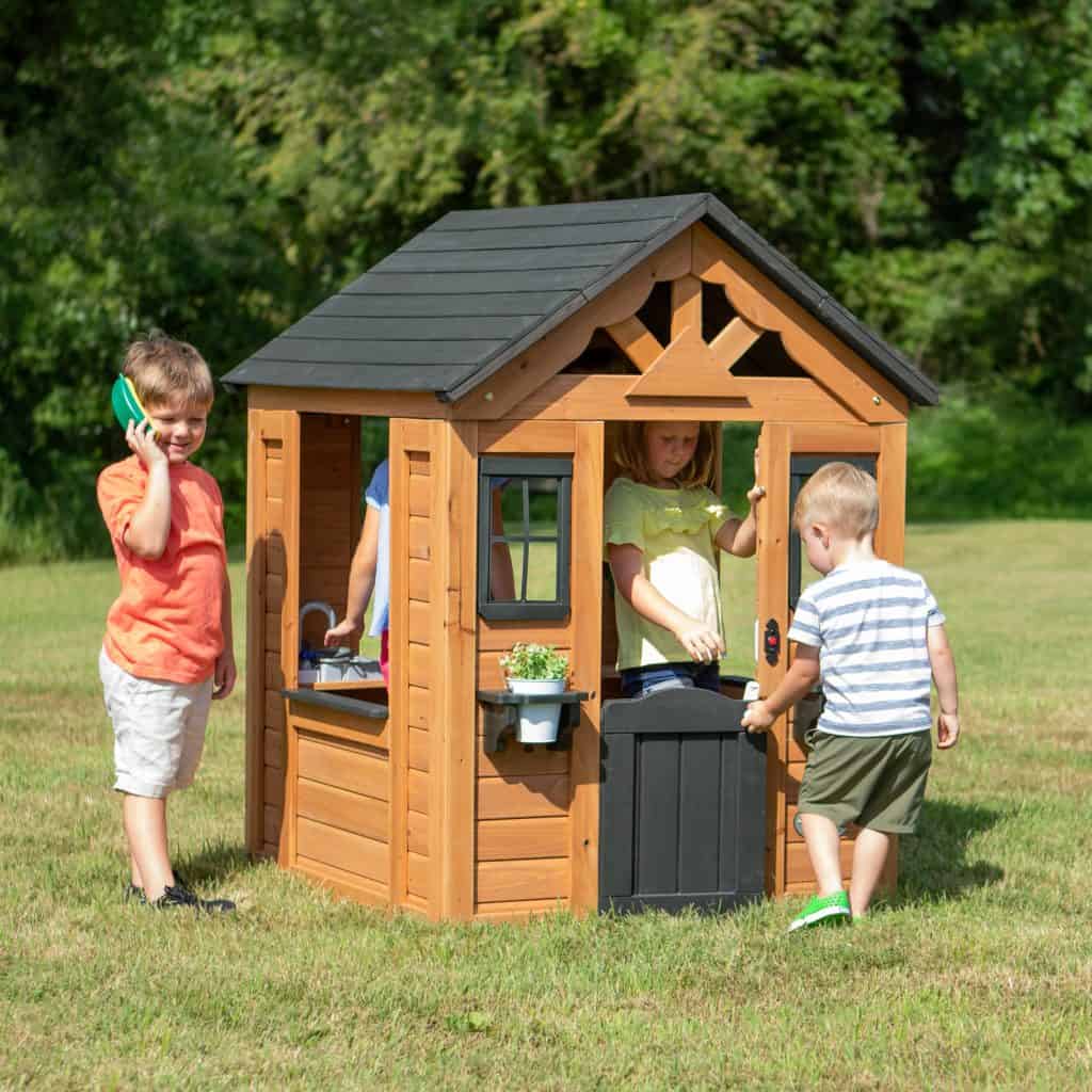 Four children are enjoying a wooden playhouse on a sunny, grassy lawn. The playhouse features a black shingled roof, a small black half-door, and a decorative sunburst gable. On the left, a boy in an orange shirt stands outside, smiling while holding a green and yellow toy phone to his ear. In the center, a young girl in a yellow top stands behind the black door, looking toward another boy in a striped shirt who is opening the door to enter. A fourth child is partially visible through the large side window, playing at the internal toy sink. The background is filled with lush green trees under bright daylight.