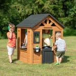 Four children are enjoying a wooden playhouse on a sunny, grassy lawn. The playhouse features a black shingled roof, a small black half-door, and a decorative sunburst gable. On the left, a boy in an orange shirt stands outside, smiling while holding a green and yellow toy phone to his ear. In the center, a young girl in a yellow top stands behind the black door, looking toward another boy in a striped shirt who is opening the door to enter. A fourth child is partially visible through the large side window, playing at the internal toy sink. The background is filled with lush green trees under bright daylight.