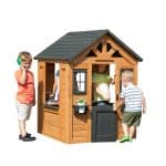 A wooden children's playhouse with a black shingled roof is isolated against a solid white background. Three children are interacting with the structure: a boy in an orange shirt stands to the left "talking" on a green toy phone, a girl in a yellow top stands inside the doorway, and a boy in a striped shirt is opening the black half-door to enter. The playhouse features a decorative sunburst gable, small windows with black frames, and a white potted plant in a window box. A toy kitchen area with a sink is visible through the large side opening of the house.