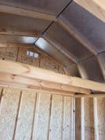 An interior eye-level shot shows the wooden framing of a shed or small building under construction. The walls and roof are made of light-colored wood studs and oriented strand board (OSB) panels. A large, horizontal wooden beam runs across the middle of the frame, likely for a loft or second floor. The roof is slanted and has a silver reflective radiant barrier on the underside. A small rectangular vent is visible on the back wall near the peak of the roof. The lighting is bright and even, suggesting it was taken during the day.