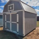 A large, gambrel-roofed storage shed, painted gray with white trim around the double doors and a decorative white star in the gable vent. The roof is corrugated metal, and the shed sits on a patch of grass and dirt under a bright sky with scattered clouds.