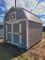 A large, gambrel-roofed storage shed, painted gray with white trim around the double doors and a decorative white star in the gable vent. The roof is corrugated metal, and the shed sits on a patch of grass and dirt under a bright sky with scattered clouds.