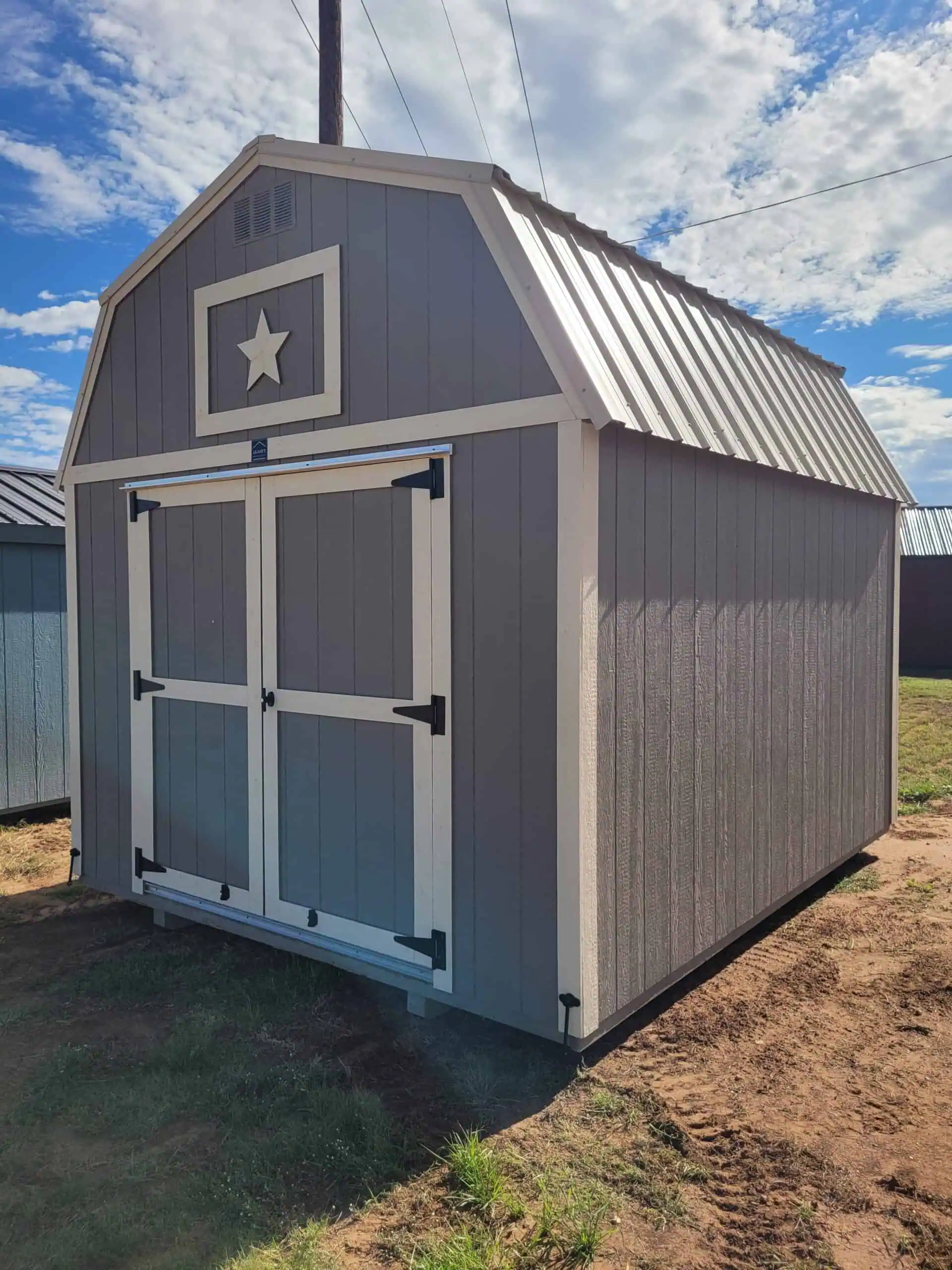 A large, gambrel-roofed storage shed, painted gray with white trim around the double doors and a decorative white star in the gable vent. The roof is corrugated metal, and the shed sits on a patch of grass and dirt under a bright sky with scattered clouds.