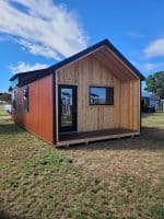 A long, modern cabin with a deep-seated front porch, featuring natural wood siding and a brick-red exterior under a blue sky.
