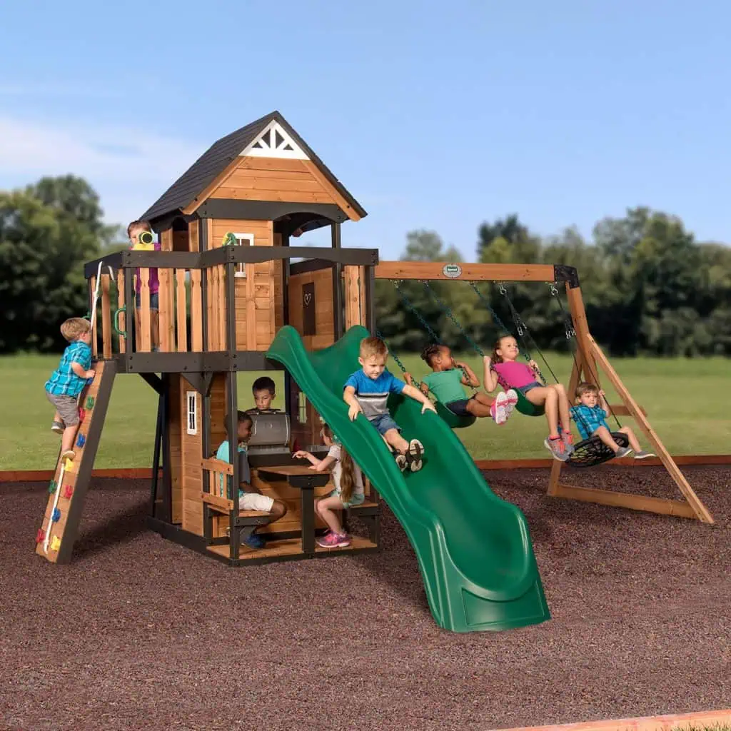 A large group of children are playing on an expansive two-story wooden playset situated on a wood-chip surface under a clear blue sky. The playset features a main tower with a black gabled roof, tan wooden siding, and black trim. On the left, a boy in a blue plaid shirt climbs a slanted rock wall leading to the upper deck, where another child peeks out. In the center, a boy in a blue t-shirt slides down a long, wavy green slide. Beneath the tower, several children are gathered at a built-in picnic table and play-kitchen area. To the right, a three-position swing beam is attached to the tower, featuring two green belt swings and a black circular webbed nest swing, all occupied by children. A lush green field and a line of soft-focus trees are visible in the background.