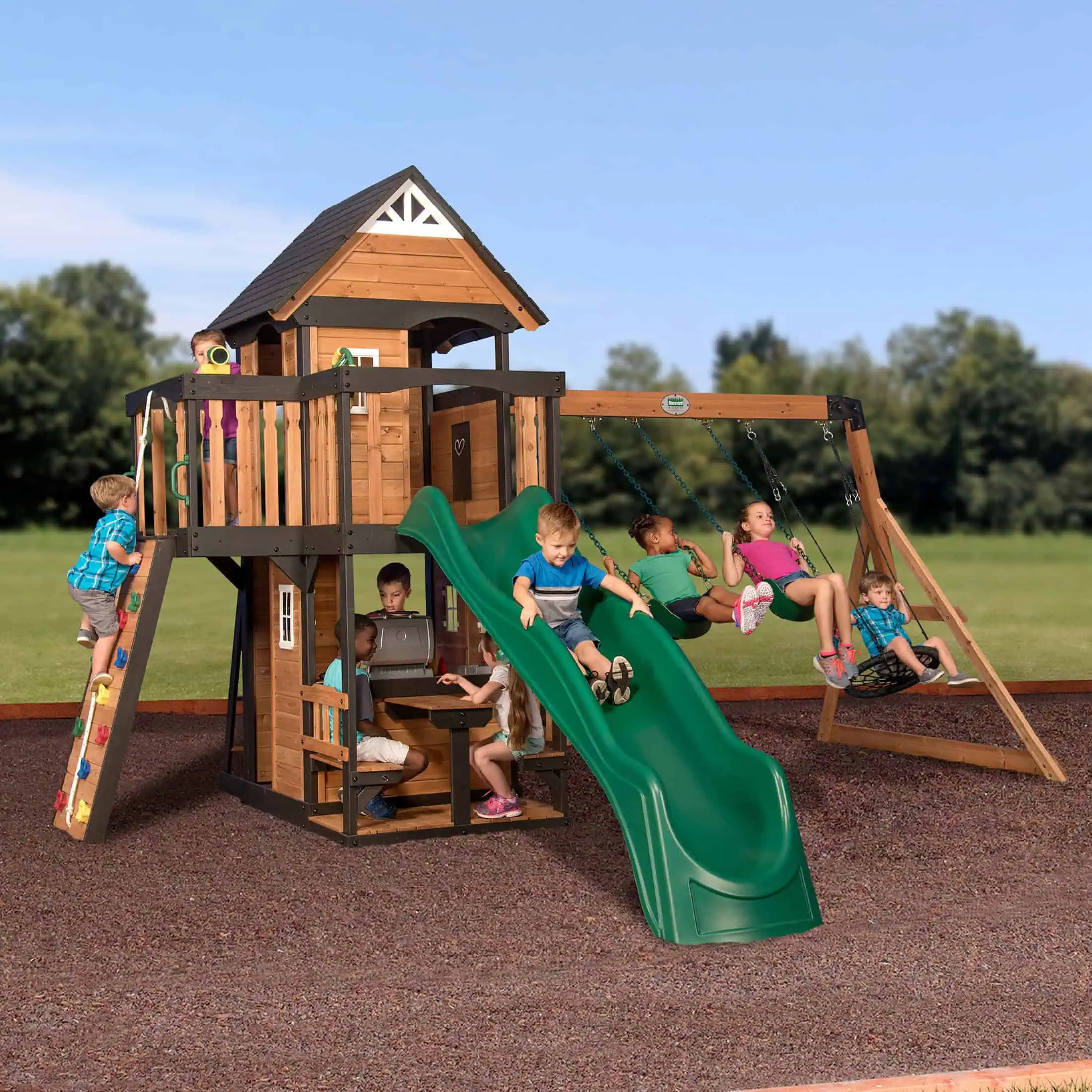 A large group of children are playing on an expansive two-story wooden playset situated on a wood-chip surface under a clear blue sky. The playset features a main tower with a black gabled roof, tan wooden siding, and black trim. On the left, a boy in a blue plaid shirt climbs a slanted rock wall leading to the upper deck, where another child peeks out. In the center, a boy in a blue t-shirt slides down a long, wavy green slide. Beneath the tower, several children are gathered at a built-in picnic table and play-kitchen area. To the right, a three-position swing beam is attached to the tower, featuring two green belt swings and a black circular webbed nest swing, all occupied by children. A lush green field and a line of soft-focus trees are visible in the background.