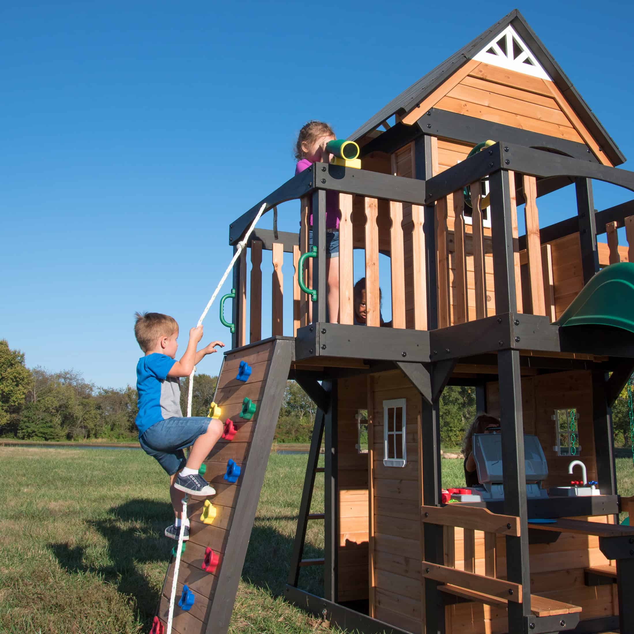 Children playing on a wooden outdoor playset in a grassy yard under clear blue sky.