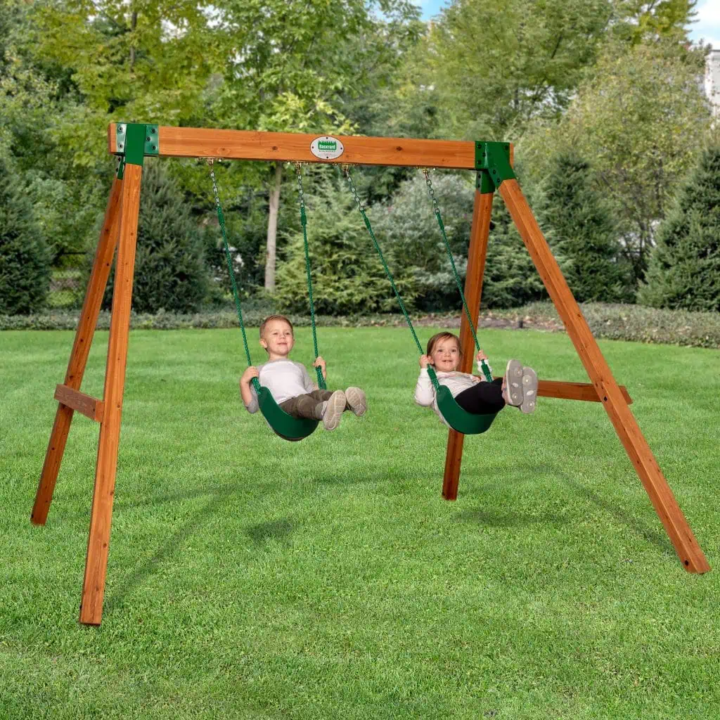 A young boy and girl are smiling while playing on a wooden A-frame swing set in a grassy yard. The swing set features a sturdy cedar wood frame with a horizontal top beam supported by angled legs and reinforced with green metal corner brackets. An oval Backyard Discovery logo is visible in the center of the top beam. Both children are seated in green flexible belt swings suspended by green ropes, set against a background of lush green trees and a clear sky.