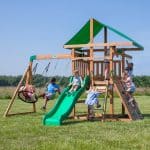 A wide shot shows a group of children playing on a large wooden playset in an open, grassy field under a bright blue sky. The playset features a tall central tower with a green fabric canopy, a wavy green slide, and a swing beam extending to the left. On the far left, a young girl swings on a black web disc swing, while a boy in a blue shirt swings on a green belt swing next to her. In the center, a boy slides down the wavy green slide, and a young girl in a blue dress climbs the ladder toward the upper deck. On the right side, a girl in a tie-dye shirt scales a wooden rock wall with green holds, while two other children are visible on the upper level—one looking through a yellow telescope and another preparing to use the rope ladder. A line of green trees is visible in the distant background.