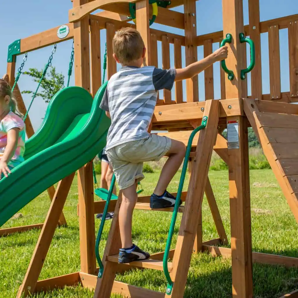 A young boy with short blonde hair, wearing a gray and black t-shirt and tan shorts, climbs a wooden ladder on a backyard playset. He holds onto a green metal handrail with his left hand and a green plastic handle on the upper deck with his right hand. To the left, a green wavy slide and a glimpse of another child are visible. The playset stands on a lush green lawn under a clear blue sky.