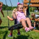 Two young girls sit back-to-back on a large, black circular web disc swing, part of a wooden backyard playset. The girl in the foreground wears a pink and white striped top, a matching pink bow in her hair, and pink sneakers, while the girl behind her has a yellow flower hair accessory. In the sun-drenched background, other children play on a vast green lawn near the wooden structure of the playset under a clear sky.