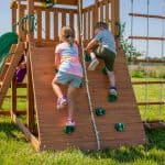 Two children, a girl in a tie-dye t-shirt and a boy in a gray t-shirt, climb up a wooden rock wall attached to a backyard playset. The wall features several green plastic climbing holds and a white knotted rope running down the center to assist them. The playset is situated on a bright green lawn under a clear sky, with a rope ladder visible to the right.