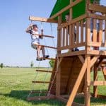 A young boy in a striped t-shirt and tan shorts climbs a white rope ladder with wooden rungs on the side of a wooden playset. Two other children watch him from the upper deck, which is shaded by a bright green canopy. The playset is situated in a wide, open grassy field under a clear blue sky.