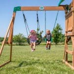 Two young girls are enjoying a sunny day on a wooden swing set in a large grassy field. On the left, a girl with a pink bow in her hair sits on a black web disc swing, looking over at her friend. To her right, another girl in a dark t-shirt smiles while swinging on a traditional green belt swing. The playset features a sturdy wooden beam with a "Backyard Discovery" logo, and the background is filled with green trees under a clear blue sky.