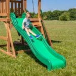 A young boy with blonde hair, wearing a light blue polo shirt, khaki shorts, and bright blue sneakers, smiles as he slides down a long, wavy green plastic slide. The slide is attached to a wooden backyard playset situated on a vast, sunlit green lawn. In the background, a line of lush green trees meets a clear blue sky.