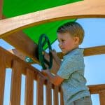 A young boy with blonde hair, wearing a gray polo shirt, stands on the wooden deck of a playset and turns a green toy steering wheel. The wheel is mounted to a wooden beam under a bright green fabric canopy. The background shows a clear blue sky, capturing a moment of imaginative play.