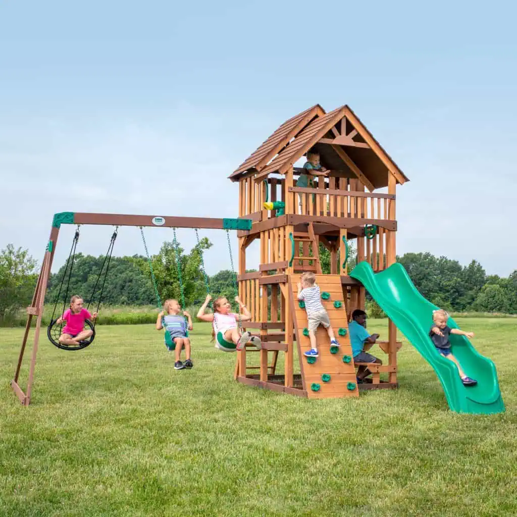 A group of seven children are actively playing on a massive, two-story wooden playset in a vast grassy field under a bright, clear sky. The wooden structure features a peaked shingle roof over the top deck, green metal safety handles, and various play components including a swing set, a green wavy slide, and a climbing wall with teal handholds.