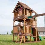 Several children are playing on a tall, three-story wooden playset in a vast grassy field under a pale sky. The top level features a wooden gable roof, and a girl in a pink shirt is looking out from the balcony. A boy is climbing a ladder toward the middle level, which is equipped with bright green safety handles. On the ground level, children are visible sitting on a built-in wooden bench. To the right, green belt swings hang from a wooden beam attached to the structure.