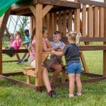 A group of children are gathered around a built-in wooden picnic table on the lower level of a backyard playset. Two children sit on one side of the table while another stands facing them, engaged in conversation. In the background, two other children are visible on swings. The structure is built from warm-toned wood and is located on a green lawn under an overcast sky.