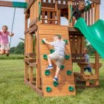 A young boy in a striped shirt and tan shorts climbs a wooden rock-climbing wall with green handholds on a backyard playset. To the left, a girl in a pink shirt is mid-air on a swing, and to the right, a girl peeks out from the top of a green wavy slide. The wooden structure features multiple levels, green safety handles, and a small picnic table area below. The scene is set on a grassy field with a line of trees in the background under a bright sky.