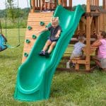A young girl with blonde hair is sliding down a bright green, wavy slide on a wooden backyard playset. Beside the slide is a wooden climbing wall with green rock-style handholds. In the background, other children are visible playing around the structure, including a boy sitting on a built-in wooden bench and two girls talking near a small picnic table area. The playset is situated on a large green lawn under a bright, overcast sky.
