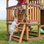 A young boy in a striped shirt and tan shorts climbs the bottom wooden ladder of a multi-level playset on a grassy lawn. Above him, a girl in a pink shirt is halfway up a second ladder leading to the highest deck, which is flanked by bright green safety handles. Other children are partially visible sitting on a lower wooden bench underneath the main platform. A green belt swing hangs from a chain on the right side of the structure.