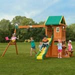 A group of children are playing on a large wooden playset situated on a grassy field under a cloudy sky. The playset features a tall tower with a green and yellow canopy, a yellow and green wavy slide, and a wooden swing beam. On the left, one child is mid-swing on a yellow belt swing, while another child stands next to a set of yellow trapeze rings. In the center, a boy is sliding down the wavy slide, and another child stands behind it. To the right, a group of children are gathered around the lower playhouse area, which includes a serving counter and an attached wooden bench. A line of dense green trees forms the background behind the play area.