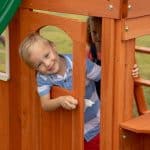 Gemini said A young, smiling boy with blonde hair and a blue-and-white striped shirt peeks through a wooden door on a playhouse. He is holding onto the white handle of the arched wooden door. In the background, partially obscured by the playhouse frame, a young girl with curly hair looks on. The playhouse is made of warm-toned wood, and a green slide is partially visible in the upper left corner.