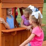 Gemini said Three young girls play together at a wooden playhouse window on a grassy lawn. One girl in a blue striped top looks out from inside the playhouse, while two other girls, one in purple and one in pink, lean against the exterior wooden counter to talk to her. The girls in purple and pink both wear large, matching white bows in their hair. The scene is captured in a close-up shot that highlights their smiling faces and the warm cedar wood of the playground structure.