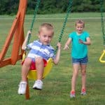 Gemini said A young boy in a blue-and-white striped shirt and red shorts sits on a yellow swing with green coated chains. He holds the chains tightly and looks toward the camera. In the background, a young girl in a teal t-shirt and denim shorts stands on the green grass, reaching out as if she just pushed the swing. The playground equipment features a sturdy wooden frame, and a wide, open green field stretches into the distance under soft daylight.