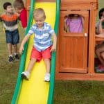 A young boy in a blue and white striped shirt and red shorts smiles as he slides down a yellow slide with green rails. He is part of a group of children playing on a wooden backyard playset situated on a green lawn. To the right, another child looks out from a window in the wooden playhouse structure, while two other children are visible on the left near a yellow swing.