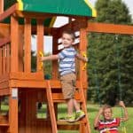 two young boys playing on a wooden swing set in a grassy backyard. One boy in a blue and white striped shirt stands on the top step of a wooden ladder, holding onto two yellow safety handles as he prepares to enter an upper-level clubhouse with a green and yellow tarp roof. Below and to the right, a second boy in a red and orange striped shirt sits on a yellow belt swing, holding the green coated chains and smiling at the camera. The playset is made of warm-toned wood, and the background features soft-focus green trees under a bright sky.