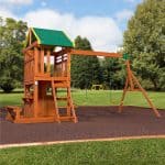 a wooden playset situated in a backyard with a green tarp roof and yellow accents. The structure features a two-story clubhouse with a lower-level play cabin and a built-in picnic bench on the side. A wooden ladder leads to the upper deck, which is enclosed by a wooden railing and has yellow safety handles. To the right, a long wooden swing beam holds two yellow belt swings and a yellow trapeze bar with rings. The playset is installed on a rectangular bed of brown rubber mulch with a wooden border, surrounded by a green lawn and a background of lush green trees under a soft blue sky.