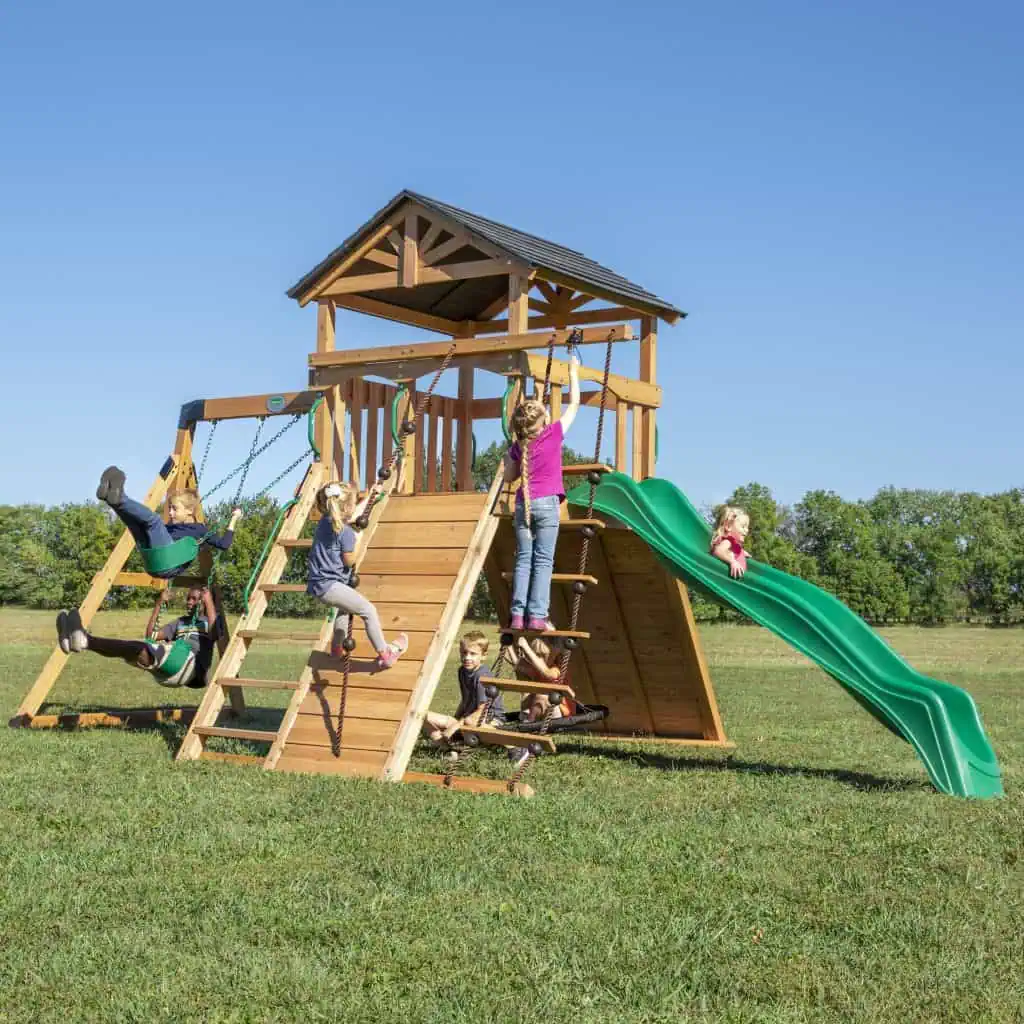 Swing Set positioned in a grassy field under a clear blue sky, with several children playing on its various components. The structure features a central elevated clubhouse with a gabled roof, accessible via a wooden ladder and a wide climbing ramp with a knotted rope. Two children are swinging on green belt swings to the left, while another child prepares to descend the long, wavy green slide on the right. Additional children are engaged in climbing a rope ladder and sitting in a web swing nestled beneath the main deck. The playset is constructed from light-toned wood with dark roof shingles and green accents, set against a background of distant green trees.