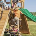 Several children are playing on a wooden outdoor playset in a grassy field on a sunny day. In the center, a young boy in an orange shirt and tan shorts climbs a rope ladder with wooden rungs. Behind him, two girls sit on a black circular web swing. To the right, a bright green wavy slide is visible, and in the background, other children are seen climbing on the upper deck of the wooden structure.