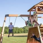 Two boys are happily swinging on a wooden swing set in an open, grassy field. The older boy on the right is swinging high, while the younger boy on the left smiles directly at the camera. In the lower right corner, another child sits on a black web swing attached to the same playset. The background features a clear blue sky and a distant line of lush green trees.