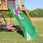 A bright, outdoor scene shows two young girls playing on a wooden playset in a grassy field. A girl in a purple shirt and glasses is halfway down a wavy green slide, while another young girl in a pink dress sits on a black circular web swing nearby. In the background, a line of lush green trees stands under a clear blue sky.