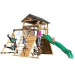 A high-angle studio shot of the Endeavor Swing Set on a white background, showing several children playing on its various features. The large wooden playset consists of a central elevated clubhouse with a dark gray shingled roof, accessible by a wooden ladder and a climbing ramp with a knotted rope. A long, wavy green slide extends to the right, while a swing beam on the left holds two green belt swings being used by two children. Other children are seen climbing a rope ladder, sitting in a black web swing underneath the deck, and playing on the main platform. The structure is made of light-toned cedar wood with green metal hardware accents throughout.
