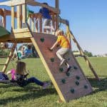 A medium-wide shot of several children playing on a large, wooden backyard playset in a sunlit, grassy field. In the foreground, a young boy in an orange shirt and tan shorts climbs a slanted wooden rock wall equipped with several green plastic handholds. To his left, a girl with blonde hair sits on a black circular web swing, while a boy in a striped blue shirt is seen mid-swing on a traditional green belt swing in the background. Another child in a blue shirt is seen moving on the upper wooden deck of the fort. The scene is set against a clear blue sky and a wide, open horizon.
