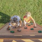 An overhead shot shows two young children, a girl with blonde pigtails and a boy in an orange shirt, climbing a slanted wooden rock wall on a playset. The wall features several green, molded plastic handholds. The children are positioned side-by-side, reaching upward as they climb toward the top. Below them, the bright green grass of a field is visible.