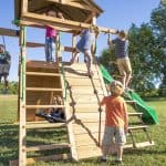 Several children are seen actively playing on a large, multi-level wooden outdoor playset in a sunlit grassy field. On the right, a young boy in a blue shirt climbs a rope ladder with wooden rungs toward the upper deck, while another boy in an orange shirt watches from below. In the center, a young girl in a blue shirt climbs a slanted wooden rock wall using a thick rope with integrated foot grips. On the left, a girl in a pink shirt stands on the upper platform under the roofed section of the fort. Below the platform, another child rests on a black circular web swing, and to the far left, a boy is partially visible swinging on a traditional green belt swing.