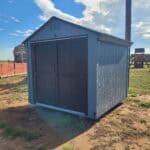 A light blue wooden storage shed with dark grey double doors sitting on a dirt and grass lot under a bright blue sky.