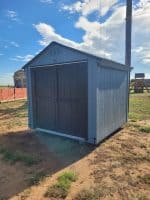 A light blue wooden storage shed with dark grey double doors sitting on a dirt and grass lot under a bright blue sky.