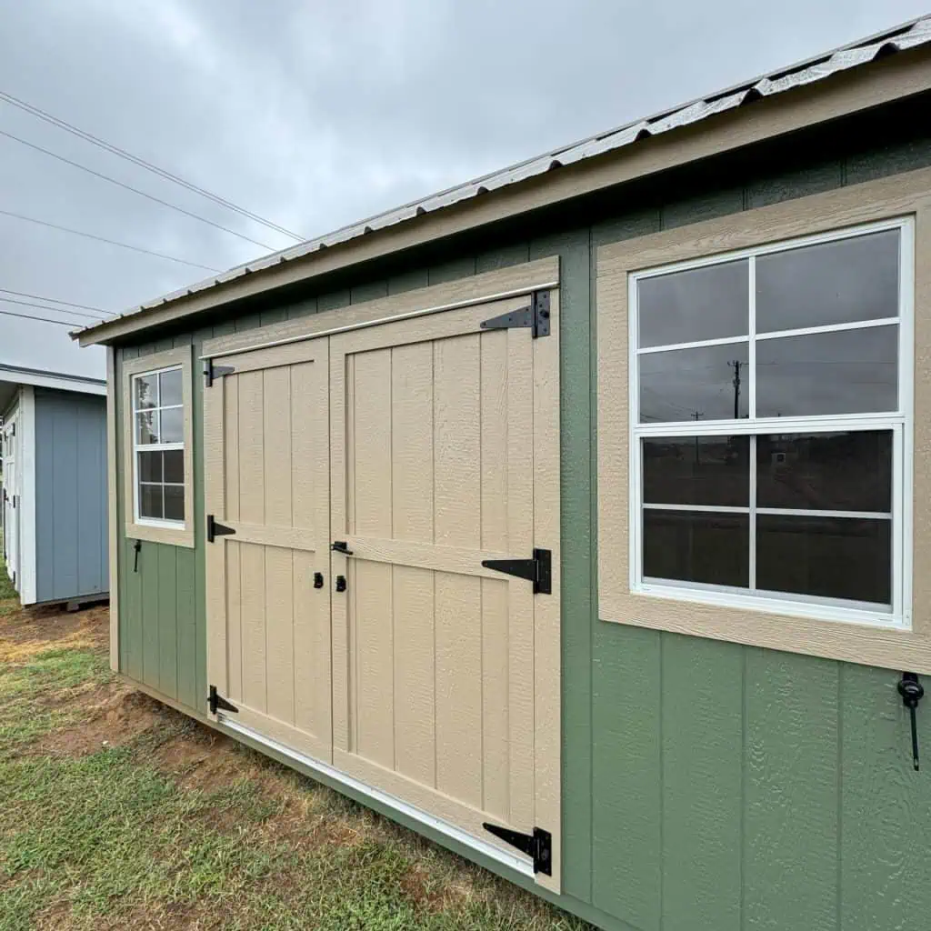 A horizontal shot of the side of a light green shed with tan trim and doors. The shed features a large window with four panes on the right, and a smaller window with two panes to the left of the tan double doors, which have black hinges and handles. The shed sits on a grassy area under a cloudy sky, with another shed partially visible in the background to the far left.