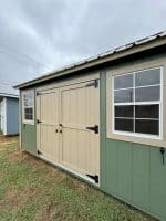 A horizontal shot of the side of a light green shed with tan trim and doors. The shed features a large window with four panes on the right, and a smaller window with two panes to the left of the tan double doors, which have black hinges and handles. The shed sits on a grassy area under a cloudy sky, with another shed partially visible in the background to the far left.