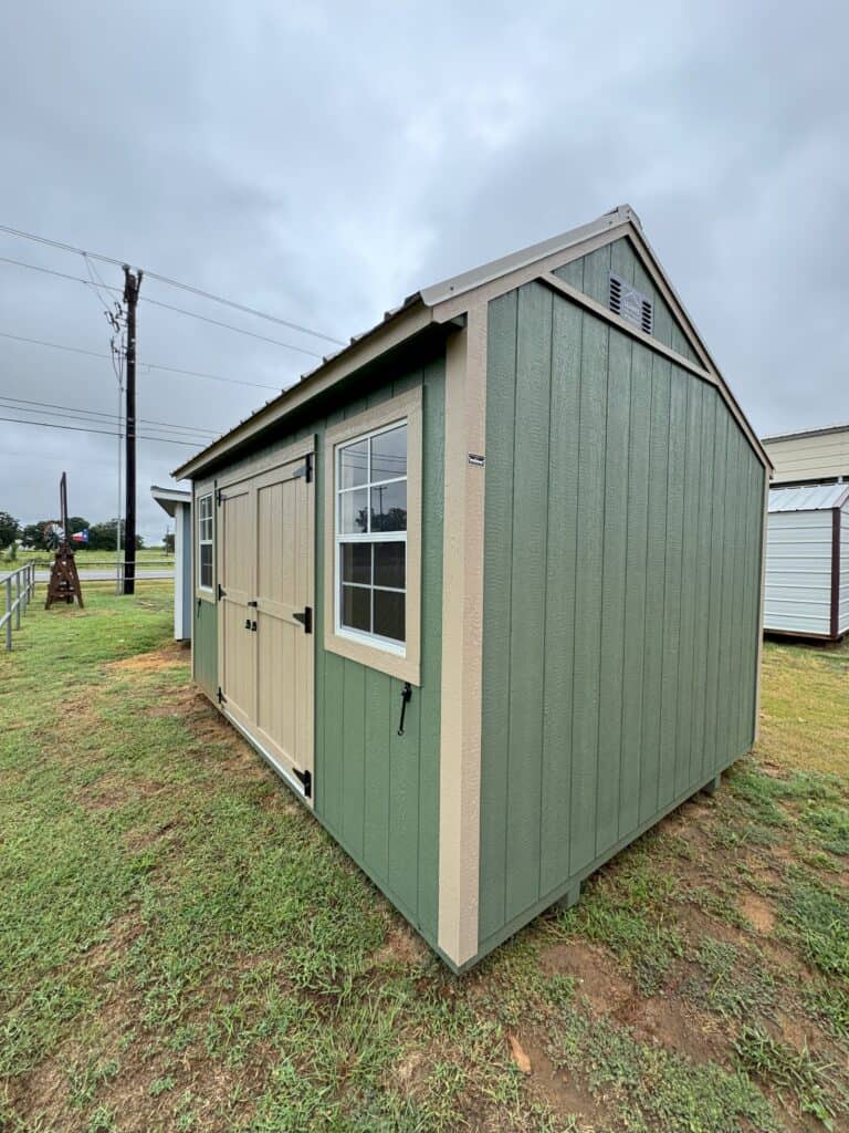 A light green wooden shed with tan trim stands on a grassy patch under a cloudy sky. The shed has a small window on its side and light-colored double doors. To the left, a fence and utility pole are visible in the background, along with another smaller shed.