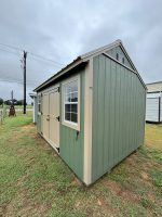 A light green wooden shed with tan trim stands on a grassy patch under a cloudy sky. The shed has a small window on its side and light-colored double doors. To the left, a fence and utility pole are visible in the background, along with another smaller shed.