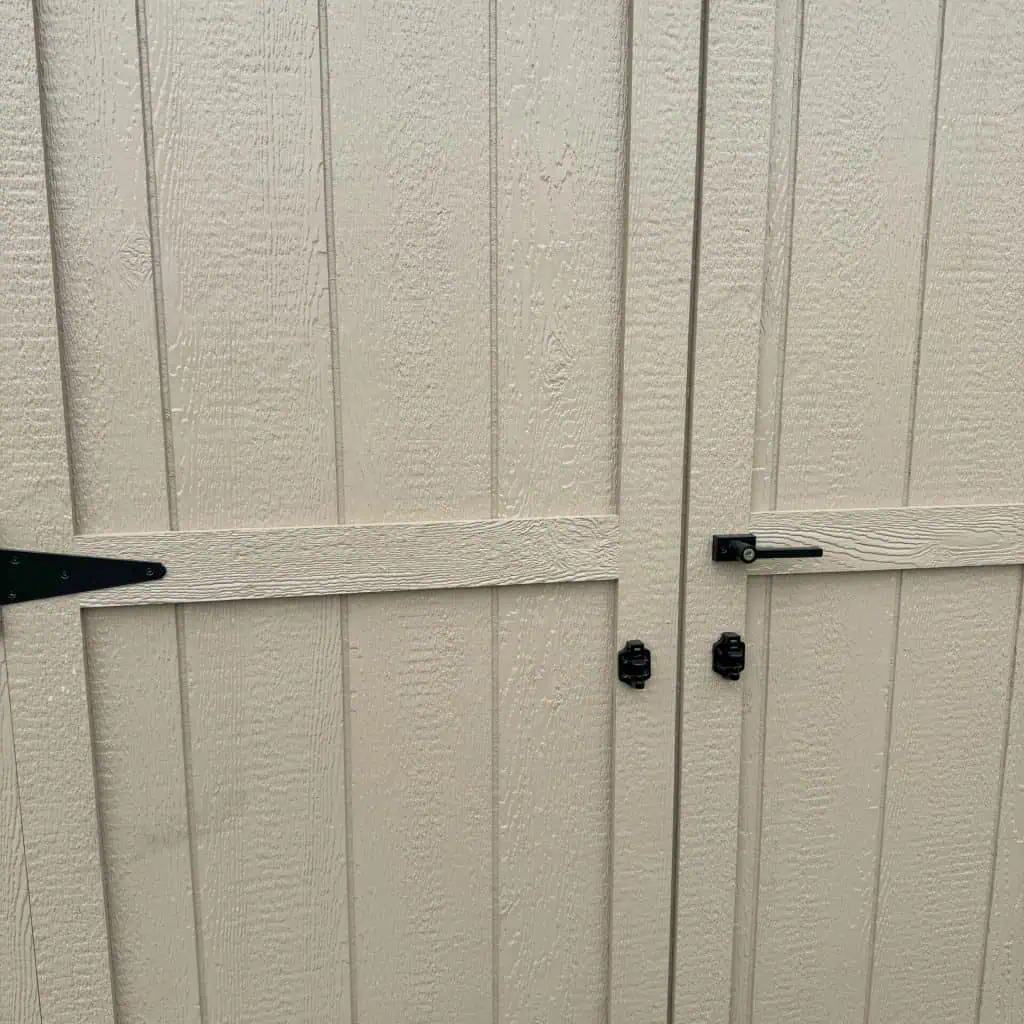 A close-up view of two light tan shed doors with a textured, wood-grain like surface. The doors have black hardware, including hinges on the left door and a latch and handle on the right door, suggesting they open outward. A seam runs vertically between the two doors.